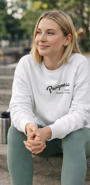 Woman wearing Progress Over Perfection white sweatshirt, sitting outdoors casual motivational streetwear