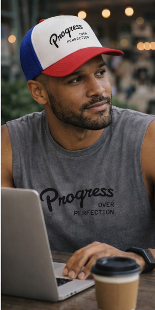 Man wearing a 'Progress Over Perfection' cap and tank top, sitting at a laptop with a blurred background.