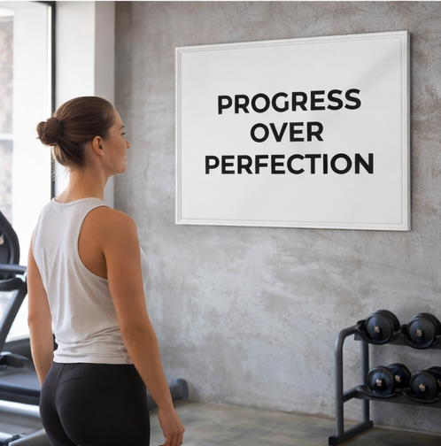Woman in a gym looking at a motivational poster with 'Progress Over Perfection' text.