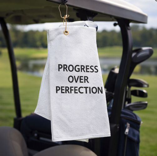 Golf towel with 'Progress Over Perfection' text hanging from a golf cart on a golf course.