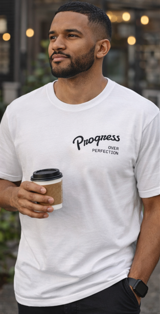 Man wearing a white unisex t-shirt with 'Progress Over Perfection' text, holding a coffee cup casual outdoor setting