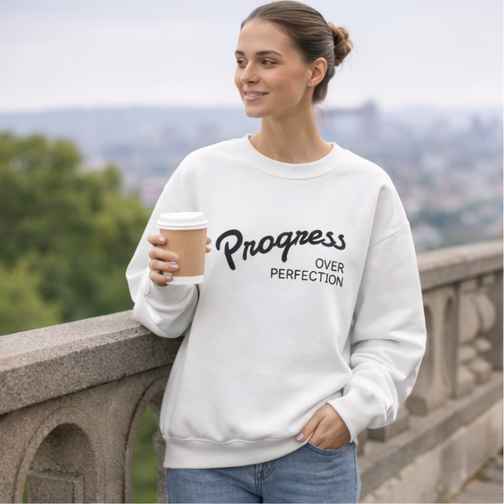 Woman wearing progress over perfection white crewneck sweatshirt outdoors with coffee, motivational apparel for mindful living and personal growth
